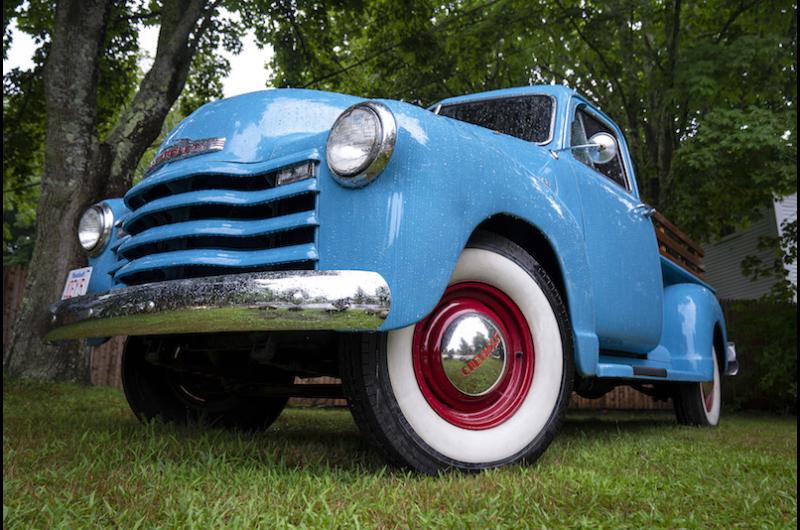 a close-up photo of the front of a baby-blue vintage pickup truck with white sidewall tires and red rims